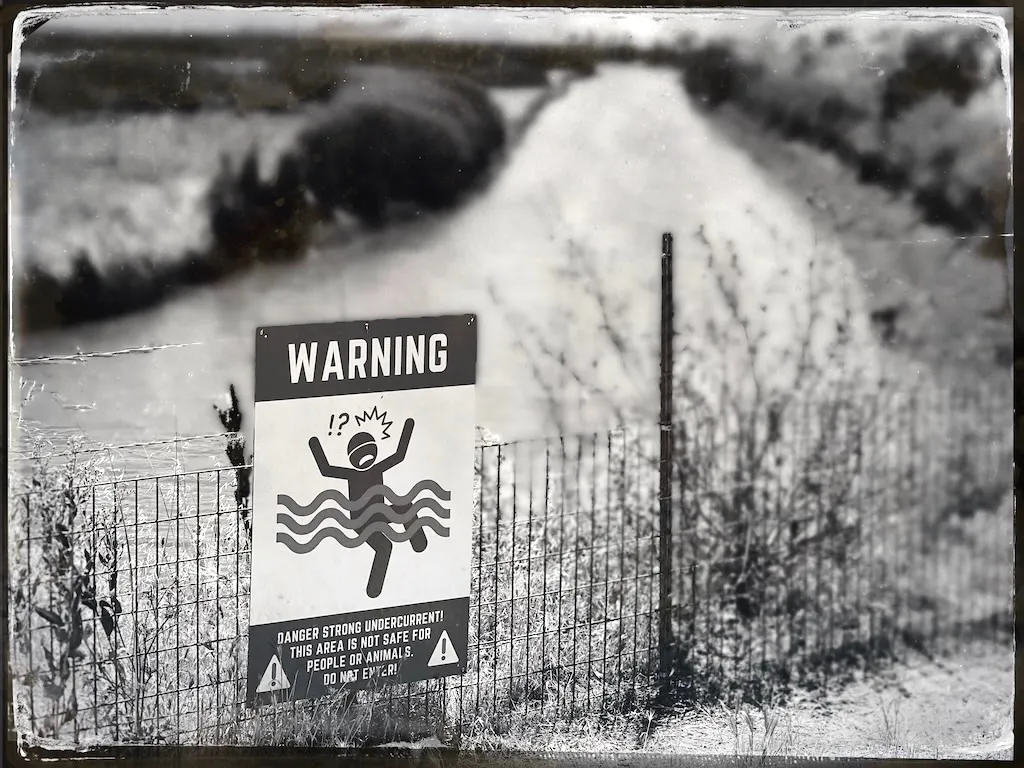 Old black and white photo of a sign on a fence at a river bend. “Danger strong current! Area not safe for people or animals.”