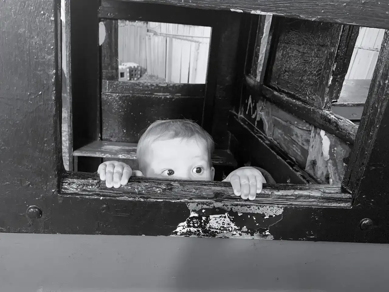 a small child's fingers and eyes peeking out from a ramshackle window