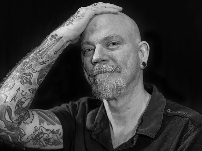 Photo of an aging white guy with a a messy goatee. His heavily tattooed right arm and hand are resting on his bald head.