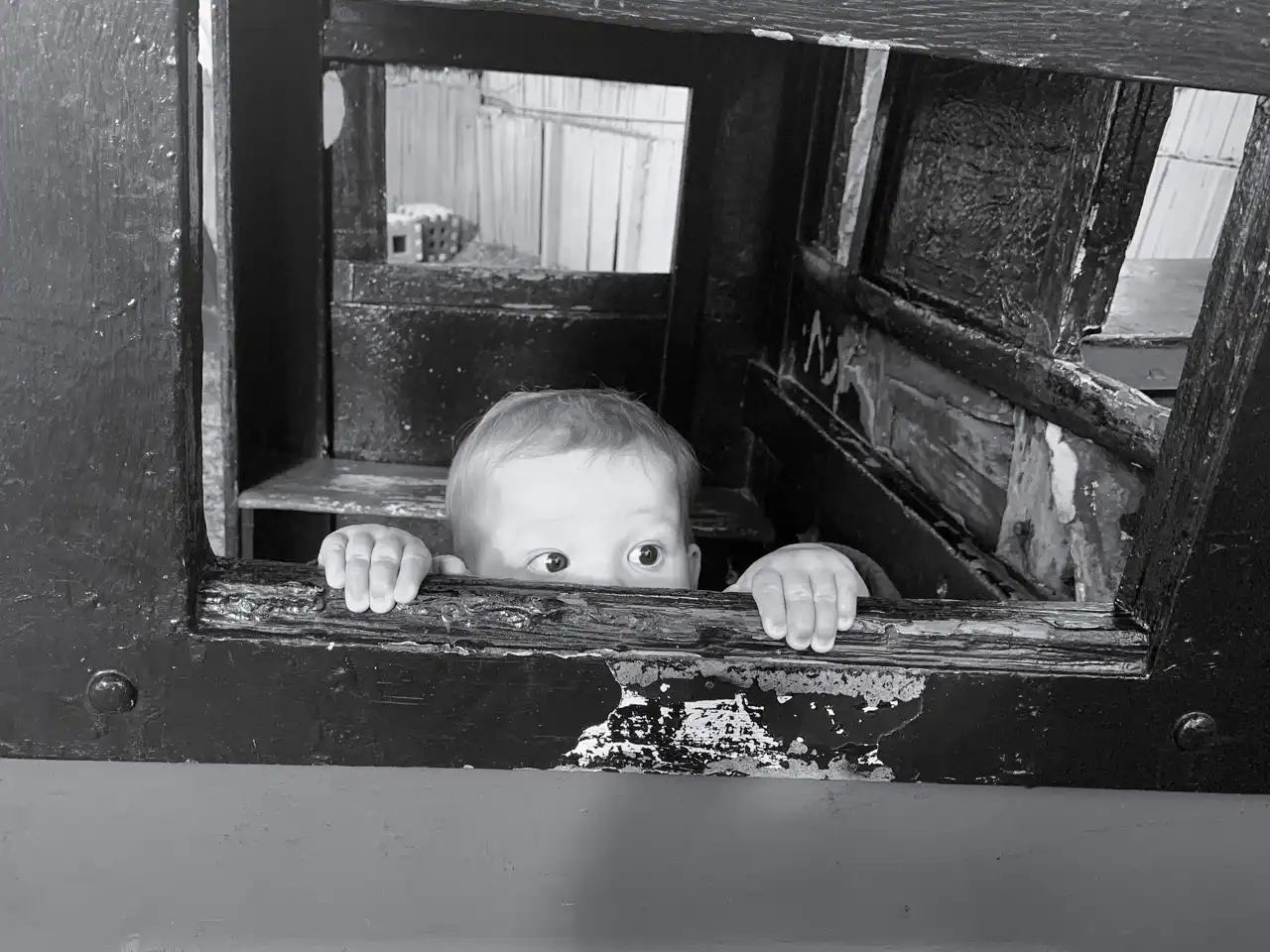 Black and white photo of large toddler eyes peeking out from a wood playhouse window, with their fingers gripping the sill.